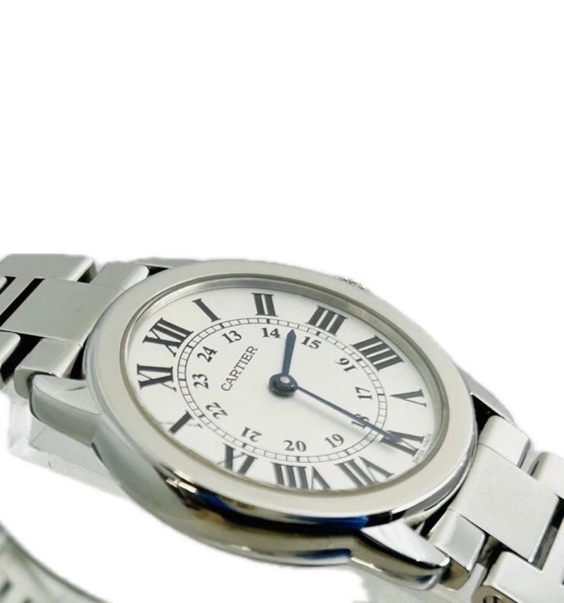 May include: A silver-toned wristwatch with a white face and black Roman numeral hour markers. The watch features a silver band and the brand name "CARTIER" is visible on the watch face. The watch hands point to the time.