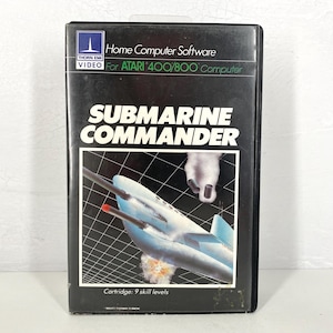 May include: Black video game cartridge for the Atari 400/800 computer. The cartridge features the game title "SUBMARINE COMMANDER" in large white letters. The cover art depicts a blue submarine with a red missile firing, set against a grid background.