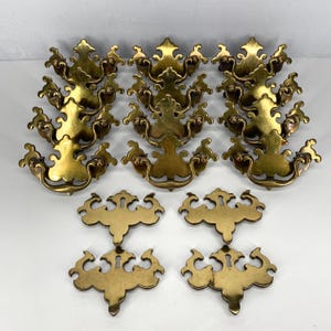 May include: A collection of antique brass drawer pulls and escutcheons. The drawer pulls have a decorative, ornate design with a curved handle. The escutcheons are keyhole-shaped with a matching design. The hardware is a warm, golden color.