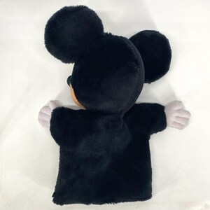 May include: A black Mickey Mouse hand puppet made of plush material. The puppet features large, round ears, a tan face, and gray hands. Designed to fit over the hand, it's a soft, fuzzy toy for play.