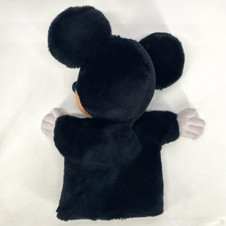 May include: A black Mickey Mouse hand puppet made of plush material. The puppet features large, round ears, a tan face, and gray hands. Designed to fit over the hand, it's a soft, fuzzy toy for play.