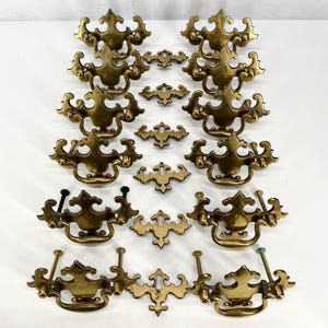 May include: A collection of antique brass drawer pulls and backplates. The drawer pulls have a decorative, ornate design with curved handles and mounting hardware. The backplates are smaller, with a matching design. The hardware is arranged on a white surface.