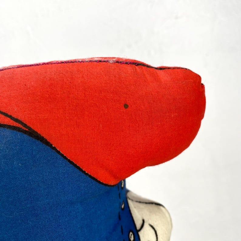 May include: Close-up of a handmade fabric doll head. The doll's head is primarily red, with a blue outfit and black outline. The doll's face is partially visible, with a white and black design.