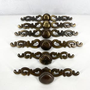 May include: A collection of antique brass drawer pulls and knobs. The pulls have an ornate, scroll-like design with a central knob. The brass finish varies in tone, from a darker bronze to a brighter gold. The knobs are oval-shaped.