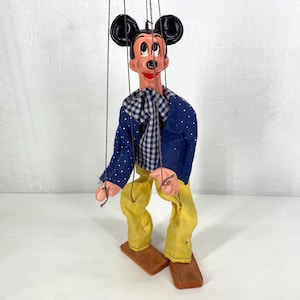 May include: Vintage Mickey Mouse marionette puppet. The puppet has black ears, a painted face, and is dressed in a blue jacket with white polka dots, a checkered shirt, and yellow pants. The puppet is attached to strings and stands on wooden blocks.