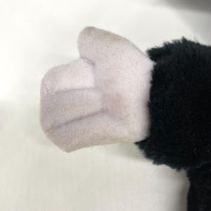 May include: A plush toy hand giving a thumbs-up. The hand is light pink, with a black furry sleeve. The toy is made of soft fabric and is likely part of a larger stuffed animal.