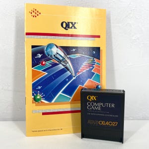 May include: A vintage QIX computer game cartridge and box. The box is yellow with red accents and a colorful graphic of a futuristic scene. The cartridge is black with white text, including "QIX COMPUTER GAME" and "ATARI CXL4027."