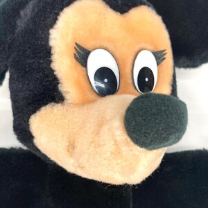 May include: Close-up of a plush toy depicting a cartoon character with a black head and ears, a tan face, and a black nose. The character has large white eyes with black pupils and black eyelashes. The toy is made of soft, fuzzy material.