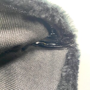 May include: Close-up of a section of dark grey fabric with a textured knit pattern. The edge is trimmed with a band of dark grey faux fur. The fabric is folded over, revealing the interior construction and stitching.