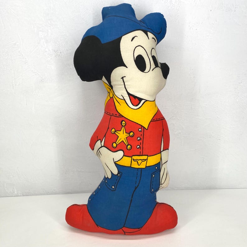 May include: A vintage Mickey Mouse plush toy in a cowboy outfit. The plush has a blue cowboy hat, red shirt with a gold star, blue jeans, and red boots. The toy has a yellow bandana and a gold belt buckle.