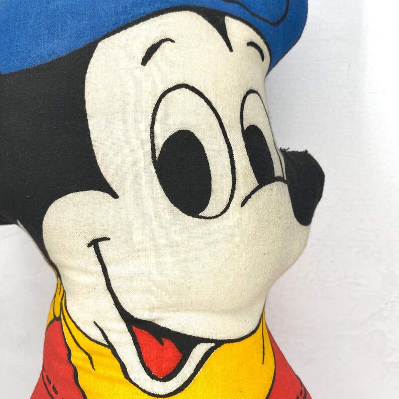May include: A vintage Mickey Mouse plush doll. The doll features a blue hat, black ears, and a red and yellow outfit. The face is white with black eyes and a smiling mouth. The plush is soft and appears well-loved.