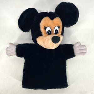 May include: A Minnie Mouse hand puppet made of plush material. The puppet has a black body, large black ears, and a tan face with black eyes, eyelashes, and a black nose. The hands are light grey. Designed to fit over the hand for play.