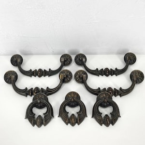 May include: A collection of six antique bronze drawer pulls and handles. The handles have a curved design with decorative floral accents. The drawer pulls have a decorative floral design with a curved handle.