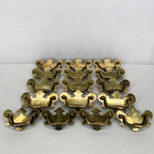May include: A collection of fifteen brass drawer pulls. Each pull features a curved handle and decorative backplate. The brass has a warm, golden hue, and the pulls are arranged in a grid pattern against a neutral background. These are suitable for furniture restoration.