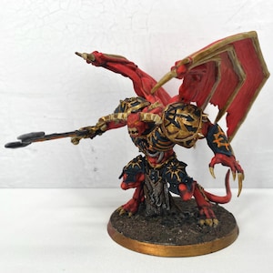 May include: A detailed miniature figure of a demonic creature with red skin, large wings, and golden armor. The figure is holding a spear and is posed on a circular base. The wings are red with gold trim.