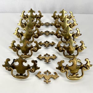 May include: A collection of antique brass drawer pulls and decorative hardware. The pulls have a curved handle design with ornate, symmetrical detailing. The hardware includes various sizes and shapes, all with a warm, aged brass finish.