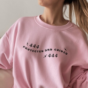 Könnte beinhalten: Rosafarbener Pullover mit dem Aufdruck "444 protected and guided 444" auf der Vorderseite.