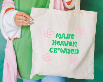 May include: A white canvas tote bag with green text that reads "Make Heaven Crowded". The text is in a retro font style. There are pink flower illustrations on the bag.