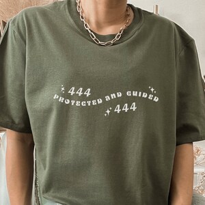 Könnte beinhalten: Ein grünes T-Shirt mit dem Text "444 Geschützt und Geleitet 444" in weißen Buchstaben. Das Shirt hat eine lockere Passform und einen Rundhalsausschnitt.