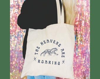 May include: Cream-colored tote bag with the text "THE HEAVENS ARE ROARING" encircling a tiger illustration in navy blue. The bag has two cream-colored straps.