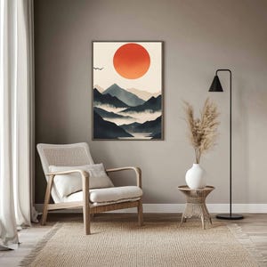 Puede incluir: Una impresión artística enmarcada con un diseño minimalista. La obra representa un gran sol naranja sobre montañas azules y grises en capas. Un pequeño pájaro vuela en el cielo. La impresión cuelga sobre una silla beige y una mesa auxiliar tejida.