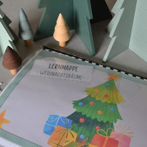 Op de afbeelding: Een groene en witte leerkaart met de titel "LERNMAPPE - WEIHNACHTSBAUME" en een kleurrijke illustratie van een kerstboom met cadeautjes eronder.