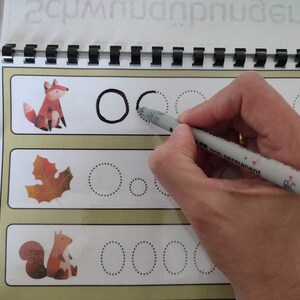 May include: A workbook with dotted lines for tracing circles. The workbook features illustrations of a fox, a leaf, and a squirrel. A hand is tracing a circle with a grey marker.