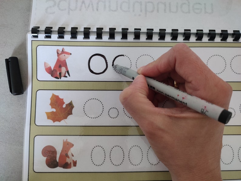 May include: A workbook with dotted lines for tracing circles. The workbook features illustrations of a fox, a leaf, and a squirrel. A hand is tracing a circle with a grey marker.