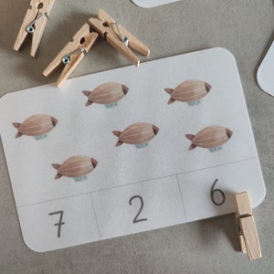 K&ouml;nnte beinhalten: Eine wei&szlig;e Karte mit sechs braunen Luftschiff-Illustrationen. Die Karte ist in drei Abschnitte unterteilt, auf denen die Zahlen 7, 2 und 6 in Schwarz geschrieben sind. Eine Holzklammer ist an der Karte befestigt.