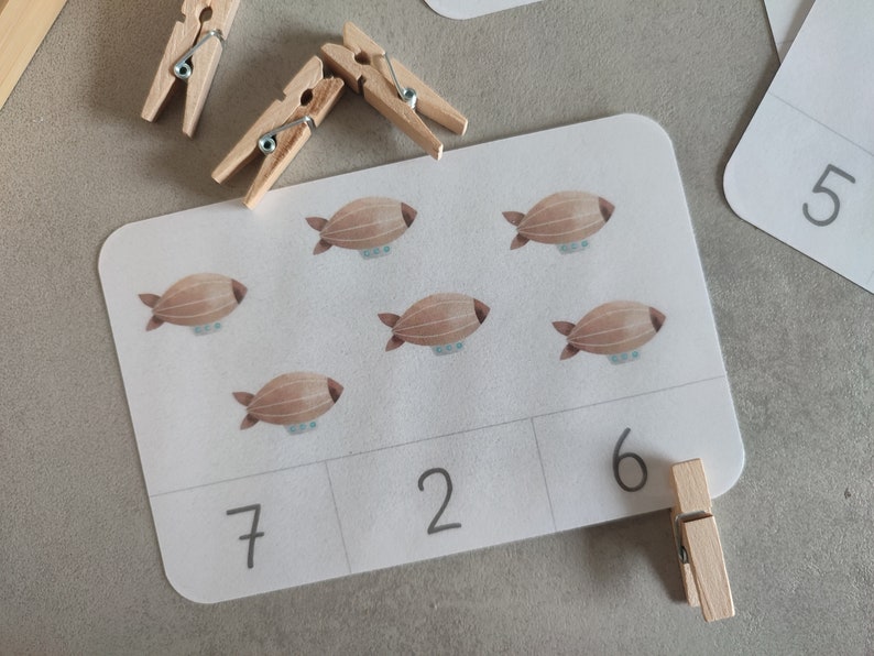 K&ouml;nnte beinhalten: Eine wei&szlig;e Karte mit sechs braunen Luftschiff-Illustrationen. Die Karte ist in drei Abschnitte unterteilt, auf denen die Zahlen 7, 2 und 6 in Schwarz geschrieben sind. Eine Holzklammer ist an der Karte befestigt.