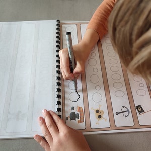 May include: A child traces dotted lines with a black marker in a workbook with pirate themed illustrations. The illustrations include a pirate ship, a skull and crossbones, an anchor, and a pirate flag.