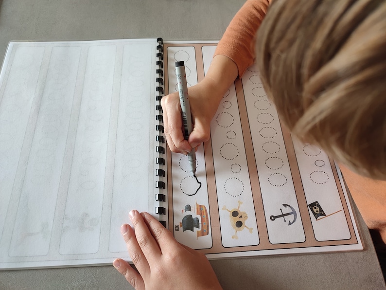 May include: A child traces dotted lines with a black marker in a workbook with pirate themed illustrations. The illustrations include a pirate ship, a skull and crossbones, an anchor, and a pirate flag.