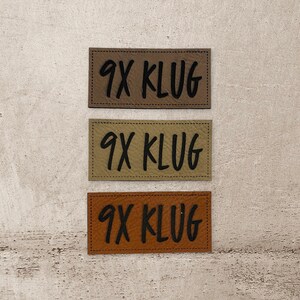 Könnte beinhalten: Drei Leder-Patches mit dem Text "9X KLUG" in schwarzer Schrift. Die Patches sind in verschiedenen Brauntönen.