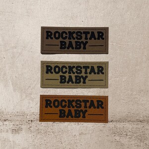 Könnte beinhalten: Drei Leder-Patches mit dem Text "Rockstar Baby" in schwarzer Schrift. Die Patches sind braun, beige und orange.
