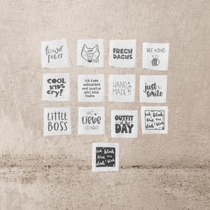 Könnte beinhalten: Eine Sammlung weißer Stoffetiketten mit schwarzem Text und Illustrationen. Die Etiketten zeigen Sätze wie "Flower Power", "Cool Kids Cry!" und "Little Boss". Weitere Etiketten zeigen eine Fuchsgrafik, eine Biene und die Worte "Hand Made".