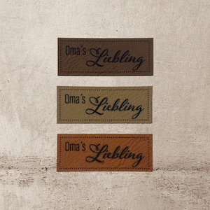Könnte beinhalten: Drei braune Leder-Etiketten mit dem Text " Oma's Liebling" in schwarzer Kursivschrift.