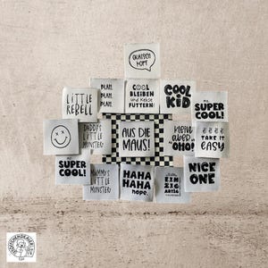 Könnte beinhalten: Eine Sammlung weißer Stoff-Etiketten mit schwarzem Text. Die Etiketten zeigen verschiedene Sätze, darunter "Cool Kid", "Super Cool", "Take It Easy", "Nice One", "Little Rebel", "Daddy's Little Monster", "Mummy's Little Monster", "Aus Die Maus", "Haha Haha Hope", "Cool Bleiben und Kekse Futtern", "Quatsch Kopf", "Klein Aber "Oho!" und "Blam. Blam. Blam."
