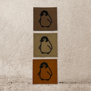 Könnte beinhalten: Drei braune Leder-Patches mit einer schwarzen Pinguin-Silhouette auf jedem. Die Patches sind vertikal angeordnet, wobei der obere Patch das dunkelste Braun, der mittlere Patch ein helleres Braun und der untere Patch das hellste Braun ist.