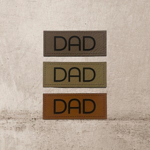 Könnte beinhalten: Drei Leder-Patches mit dem Wort "DAD" in schwarzer Schrift. Die Patches sind in verschiedenen Brauntönen.