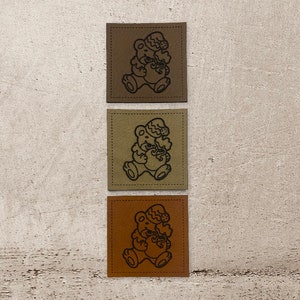 Könnte beinhalten: Drei Leder-Patches mit braunem, beigefarbenem und orangefarbenem Hintergrund. Jeder Patch zeigt eine schwarze Umrandung eines Bären, der einen Weihnachtsmann-Hut trägt und ein Geschenk hält.