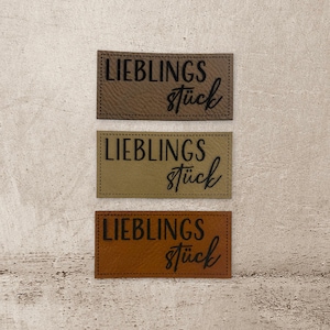 Könnte beinhalten: Drei Leder-Patches mit dem Text "Lieblingsstück" in schwarzer Schrift. Die Patches sind braun, beige und orange.