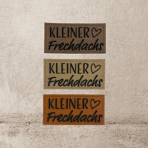 Könnte beinhalten: Drei braune Lederpatches mit dem Text "KLEINER♥ Frechdachs" in schwarzer Schrift.