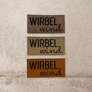 Könnte beinhalten: Drei Leder-Patches mit dem Text "Wirbel wind" in schwarzer Schrift. Die Patches sind in verschiedenen Brauntönen: hellbraun, beige und dunkelbraun.