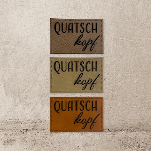 Könnte beinhalten: Drei Leder-Patches mit dem Text "Quatsch kopf" in schwarzer Schrift. Die Patches sind braun, beige und orange.