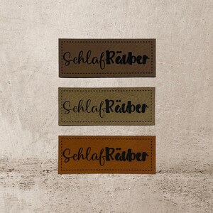 Könnte beinhalten: Drei braune Leder-Etiketten mit dem Text "Schlaf Räuber" in schwarzer Schrift.