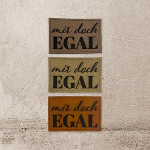 Könnte beinhalten: Drei braune Leder-Patches mit dem Text "mir doch EGAL" in schwarzer Schrift.