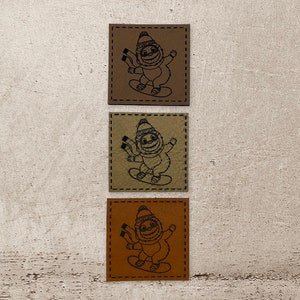 Könnte beinhalten: Drei Leder-Patches mit einem Cartoon-Faultier, das einen Hut und einen Schal trägt und Snowboard fährt. Die Patches sind braun, beige und orange.