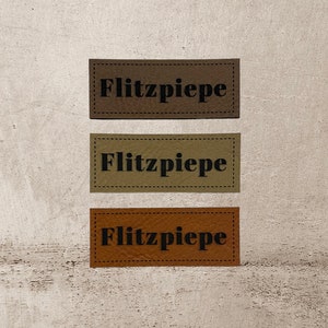 Könnte beinhalten: Drei Leder-Etiketten mit dem Text "Flitzpiepe" in schwarzer Schrift. Die Etiketten sind braun, beige und orange.