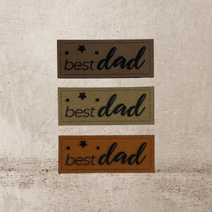 Könnte beinhalten: Drei braune Leder-Patches mit dem Text "best dad" in schwarzer Schrift und kleinen Sternen über dem Text. Die Patches sind vertikal angeordnet, wobei der obere Patch der dunkelste ist, der mittlere Patch etwas heller ist und der untere Patch der hellste ist.