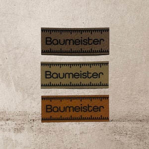 Könnte beinhalten: Drei braune Leder-Etiketten mit schwarzem Text und einem Lineal-Design. Der Text auf jedem Etikett lautet "Baumeister".